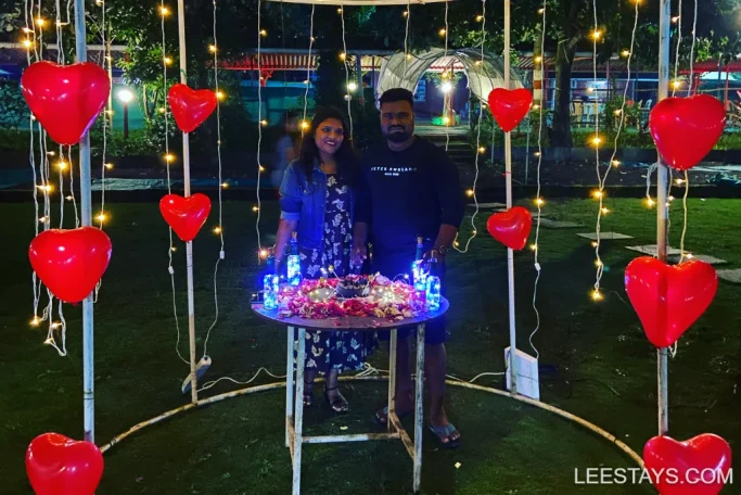 A couple stands by a decorated table surrounded by heart-shaped balloons and string lights at Lakeview Cottages in Pawna, with a lawn and illuminated area in the background.