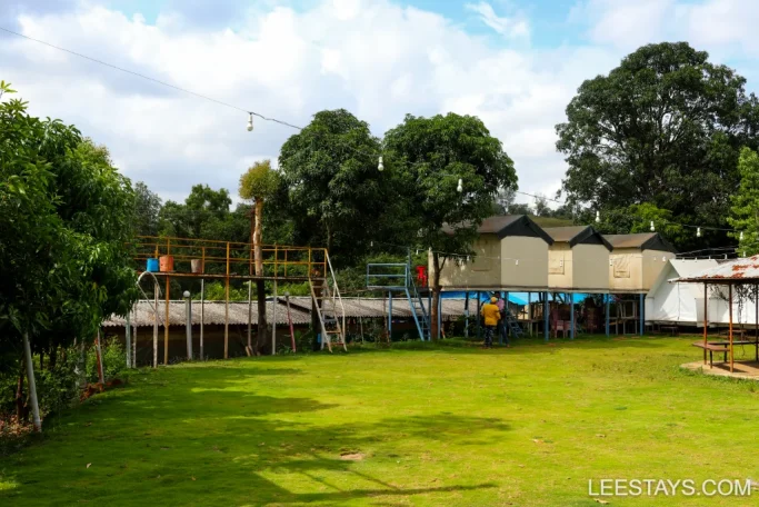 A view of raised lakeview cottages surrounded by greenery at a resort near Pawna, with a well-maintained lawn and cloudy skies overhead.