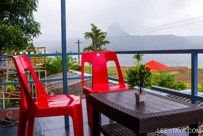 A scenic view from a lakeview cottage balcony in Pawna, featuring two red chairs, a table with a small plant, and a view of the lake and mountains in the background.
