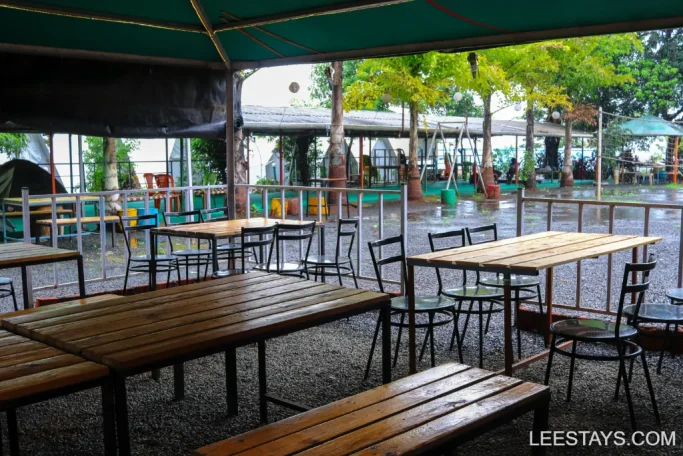 Outdoor seating area with wooden tables and chairs under a canopy, overlooking lakeview cottages in Pawna.