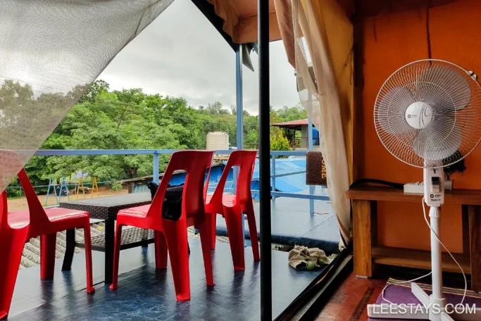 View from a lakeview cottage at Pawna, featuring a patio with red plastic chairs, a small wooden table with a fan, and lush greenery in the background.