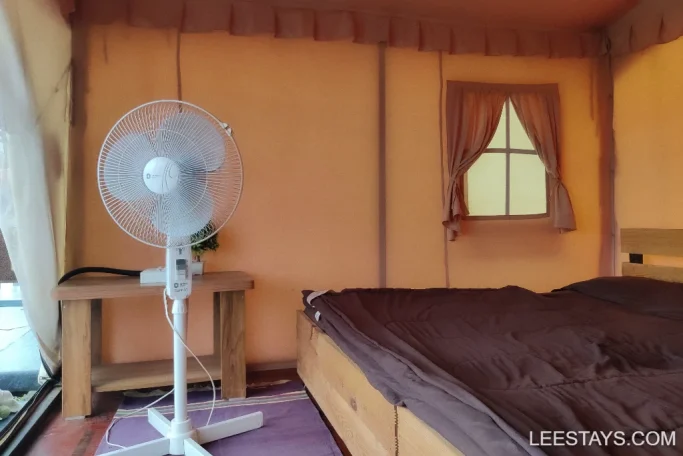Interior of a cozy Pawna lakeview cottage with a bed, bedside table, and a standing fan, featuring neutral-toned curtains and bedding.
