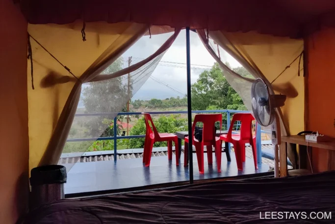 View of a scenic landscape from inside a lakeview cottage in Pawna, featuring red plastic chairs on a balcony and lush greenery outside.