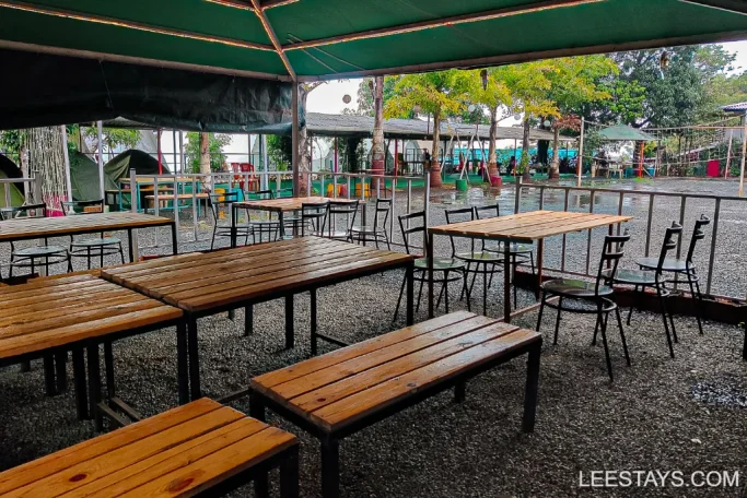 Outdoor seating area with wooden tables at Lakeview Cottages in Pawna, surrounded by trees and tents in the background.
