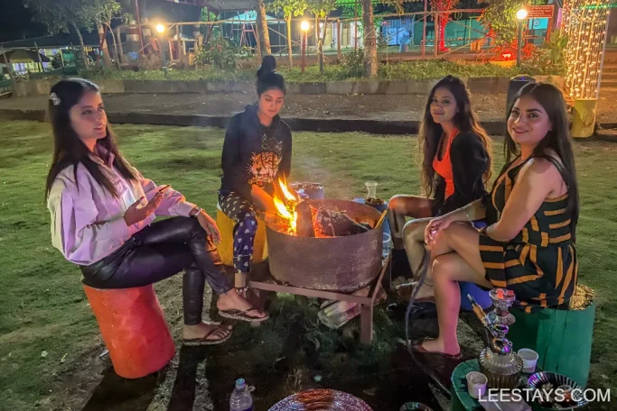 Four people sitting around a campfire at lakeview cottages in Pawna, enjoying the evening with drinks and a hookah on the grass.
