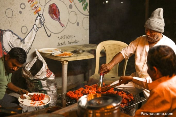 Men grilling food at a roadside stall near Pawna Lake stay, with colorful wall art in the background.