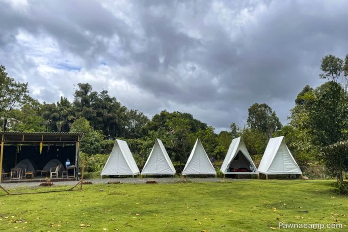 Tented stay near Pawna Lake featuring multiple white canopy tents on a grassy area, surrounded by lush greenery under a cloudy sky.