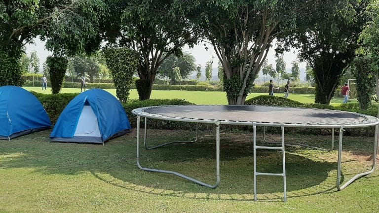 Camping area at Pawna Lake featuring two blue tents and a large trampoline set on a grassy lawn with trees offering shade.