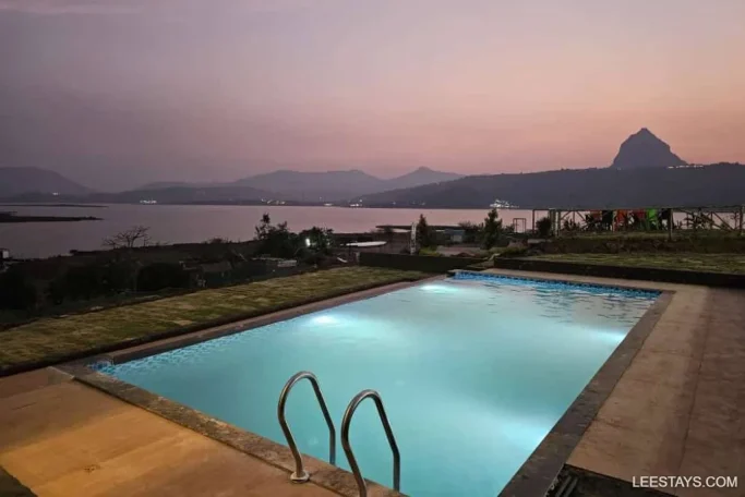 Scenic view of a lakeside retreat in Pawna with a lit swimming pool, surrounded by grass, overlooking the tranquil lake and mountainous landscape at dusk.