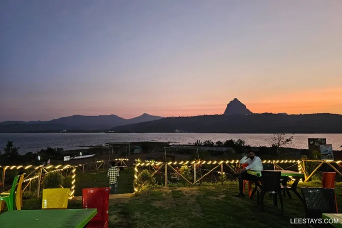 A serene lakeside retreat in Pawna at dusk, featuring a silhouetted mountain backdrop and outdoor seating adorned with lights.
