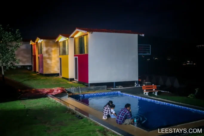 A family sits by a illuminated pool at night, beside colorful cabins at a lakeside retreat in Pawna.