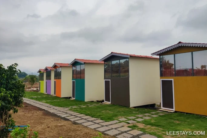 A row of colorful, modern cabins at a lakeside retreat in Pawna, with a paved path leading to them, set against a backdrop of a cloudy sky and greenery.