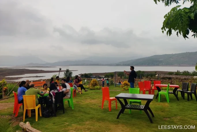 A group enjoys a meal at a colorful lakeside retreat in Pawna, with tables and chairs set on lush grass overlooking the scenic water and distant hills.