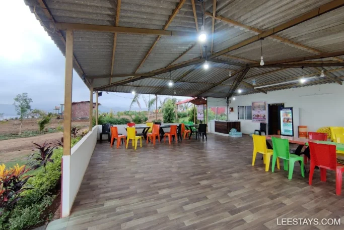 Open-air lakeside retreat in Pawna featuring colorful chairs under a metal roof, surrounded by greenery and scenic views.