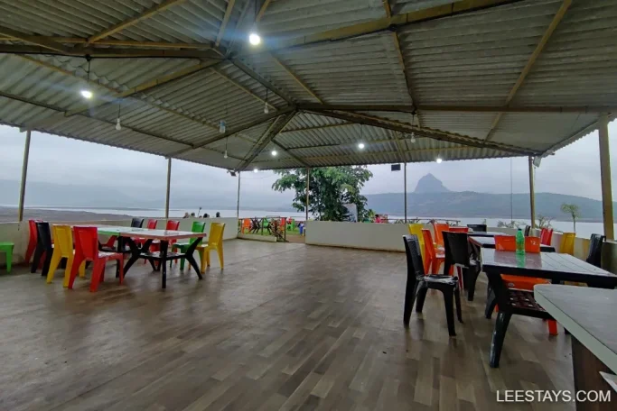 Colorful chairs and tables under a roof at a lakeside retreat in Pawna, with views of hills and misty sky.
