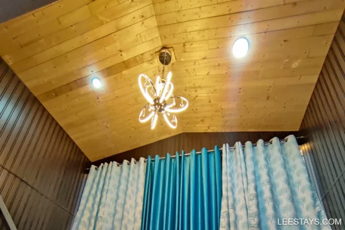 A wooden ceiling with a modern chandelier and blue curtains in a cozy room, suggesting a relaxing lakeside retreat ambiance at Pawna.