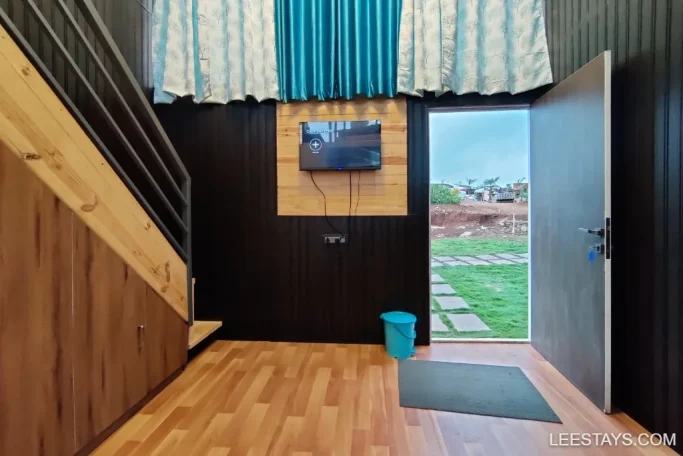 Interior of a cozy lakeside retreat near Pawna with wooden flooring, a staircase, a mounted TV, and an open door revealing a grassy outdoor area.