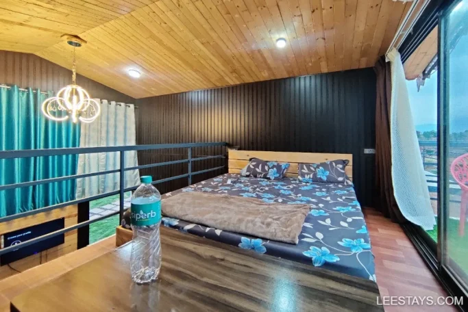 Lakeside retreat in Pawna featuring a cozy bedroom with wood paneling, floral bedspread, pendant light, and large windows overlooking a scenic view.