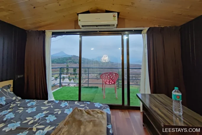 Cozy lakeside retreat room in Pawna with a bed, dresser, and a view of the lake and mountains from a large window, featuring a patio with a red chair.