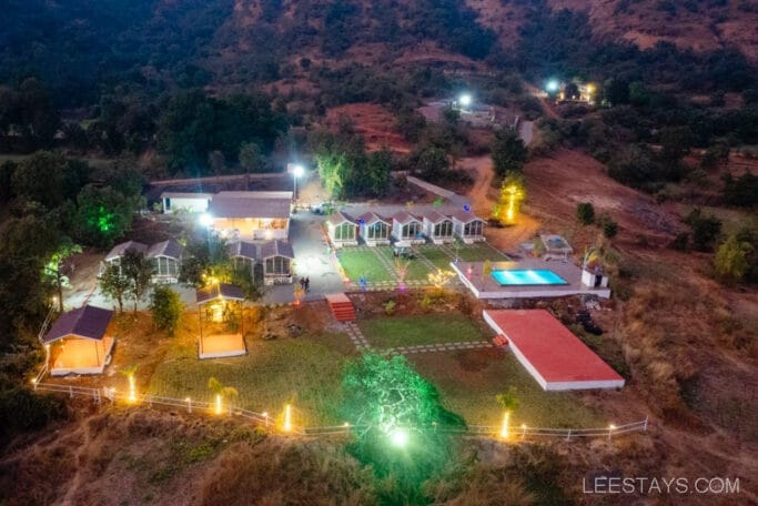 Aerial view of a budget stay near Pawna Lake with cabins, a swimming pool, and illuminated pathways surrounded by lush greenery and hills.