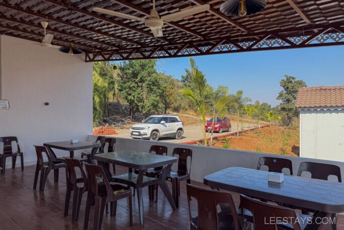 Outdoor dining area at a budget stay near Pawna Lake, featuring plastic chairs, tables, ceiling fans, and a view of parked cars and foliage.
