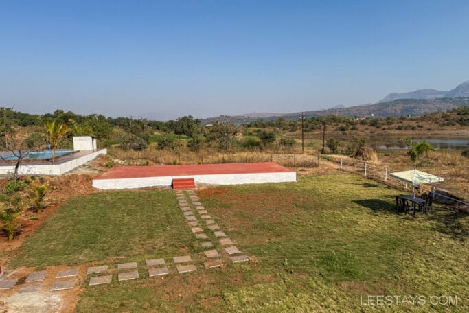 Scenic view of a budget stay near Pawna Lake, featuring an outdoor area with a pool, red platform, stone path, table with umbrella, and distant hills.