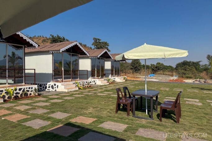 Budget stay cabins near Pawna Lake with outdoor seating under umbrella on grassy area, surrounded by scenic hills and clear sky.