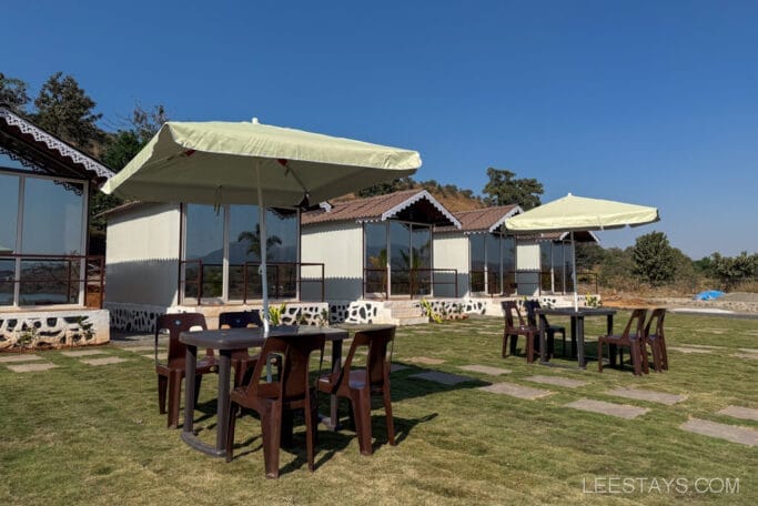 Charming cabins with glass fronts and outdoor seating under umbrellas, perfect for a budget stay near Pawna Lake, nestled in a scenic landscape.