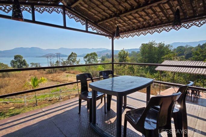 A budget stay with a view of Pawna Lake featuring a patio table and chairs overlooking scenic hills and water under a rustic roof during daytime.