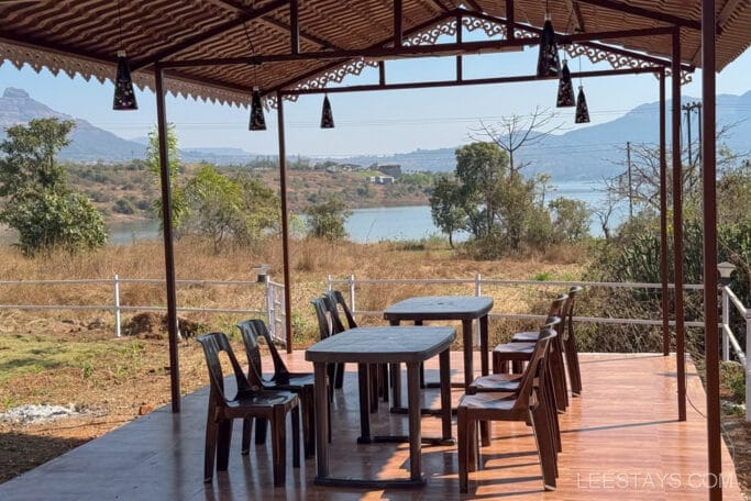 Outdoor seating area at a budget stay near Pawna Lake, featuring plastic chairs and tables under a wooden roof with scenic lake and mountain views.