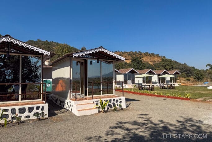Row of small cabins with glass fronts in a scenic area, ideal for budget stay near Pawna Lake, with hills and greenery in the background.