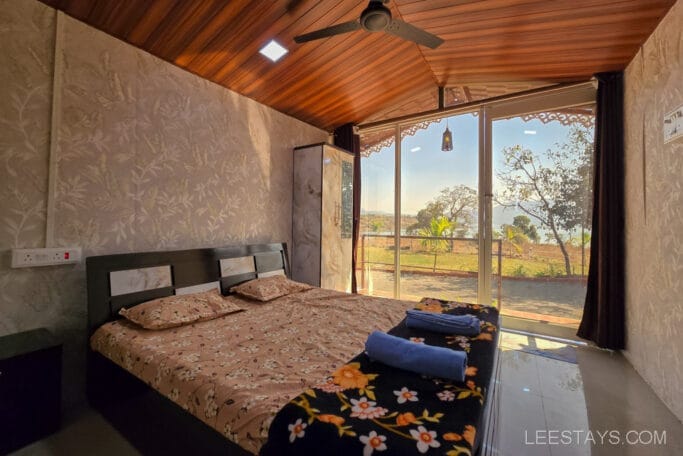 A cozy, budget-friendly room with a floral bedspread and a scenic view of Pawna Lake through a large window, offering an affordable stay near nature.