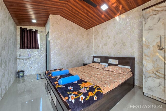 A cozy bedroom near Pawna lake, a wooden ceiling, and a bed featuring floral bedding. Two blue towels are neatly placed on the bed.