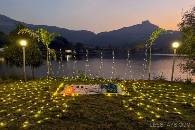A scenic view at sunset of Domestory Resort by Malvandi Lake, featuring a grassy area adorned with glowing string lights and two lamp posts, with mountains in the background.