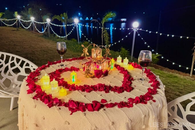 Romantic dining setup at Domestory Resort, Malvandi Lake, featuring a table decorated with red rose petals, candle holders, and two glasses of wine overlooking the water.