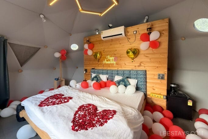 A cozy room at Domestory Resort near Malvandi Lake, decorated with red and white balloons and heart-shaped rose petals on the bed, celebrating an anniversary.