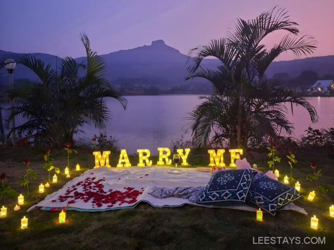 Romantic lakeside proposal setup at Domestory Resort, Malvandi Lake, featuring illuminated 'Marry Me' letters, candles, rose petals, and cushions at sunset, with a mountain backdrop.