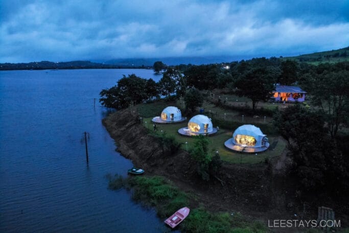 A scenic view of uniquely shaped accommodations at Domestory Resort, Malvandi Lake, featuring illuminated dome structures beside a vast lake, surrounded by lush greenery and a serene atmosphere.