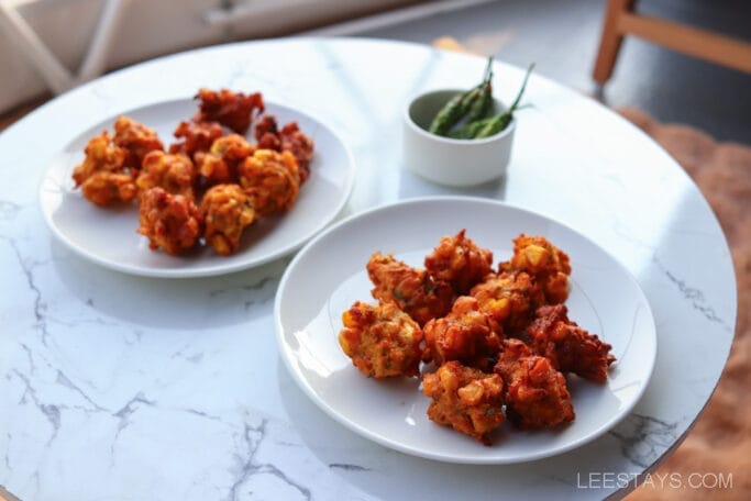 Plates of crispy fried snacks, likely served at Domestory Resort near Malvandi Lake, rest on a marble table alongside a small bowl of green chilies.