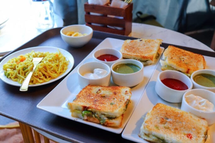 A meal setup at Domestory Resort, Malvandi Lake, featuring grilled sandwiches with dips on one plate and a serving of noodles on another, accompanied by small bowls of sides.