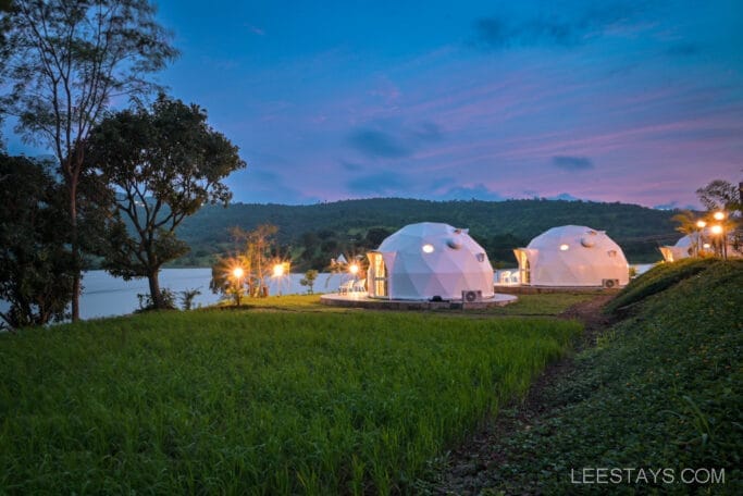 Dome-style accommodations at Domestory Resort near Malvandi Lake, surrounded by green grass and trees, with hills in the background under a colorful evening sky.