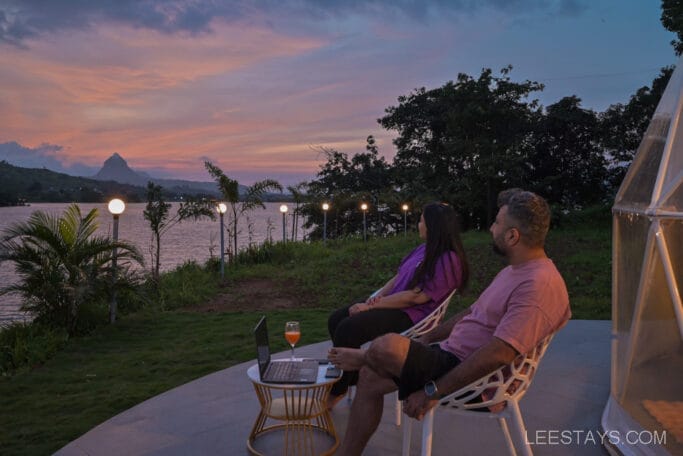 A man and woman relax at Domestory Resort, Malvandi Lake, enjoying a scenic sunset view with lush greenery, a laptop, and a drink on a table.