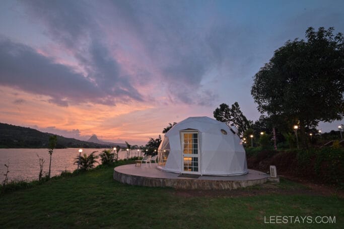 A geodesic dome at Domestory Resort, Malvandi Lake, with a scenic sunset backdrop, surrounded by greenery and illuminated by outdoor lights along the shoreline.
