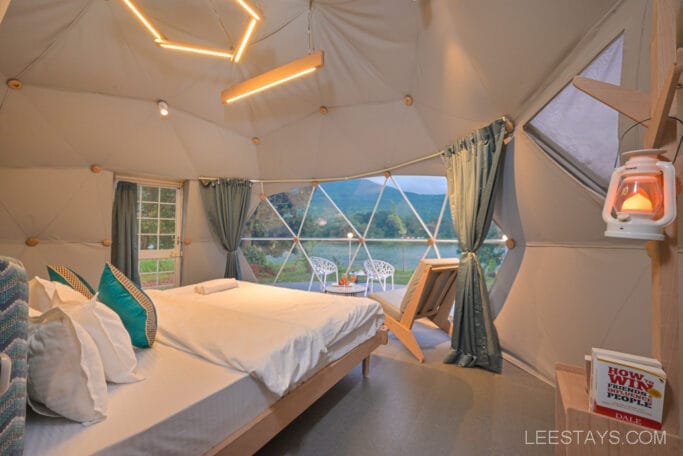 Interior of a geodesic dome room at Domestory Resort, Malvandi Lake, featuring a cozy bed, modern lighting, and a large window offering a scenic view of the lake and mountains.