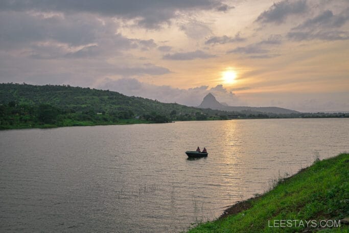 Scenic view of Malvandi Lake near Domestory Resort at sunset, featuring a small boat with two people on the calm water and lush green hills in the background.