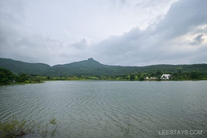 View of Malvandi lake with lush greenery, hills in the background, and Domestory Resort nestled on the shoreline under a cloudy sky.