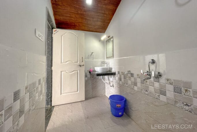 Interior of a bathroom at riverside cottages near Pawna Lake, featuring a white door, a sink with a mirror, tiled walls, and a blue bucket on the floor.