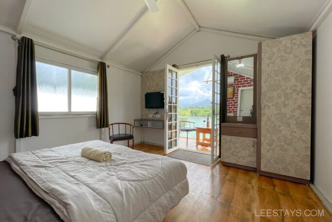 Interior of a riverside cottage near Pawna Lake, featuring a cozy bedroom with a large bed, wooden flooring, and a view through French doors that open to a deck with a scenic lake background.