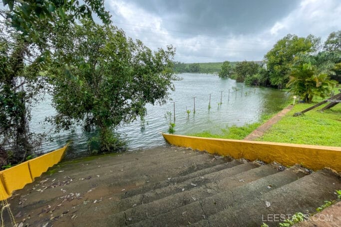 View of Pawna Lake with riverside cottages nearby, featuring a staircase leading down to the water and surrounded by lush greenery and trees on a cloudy day.