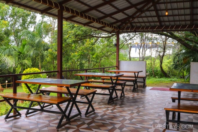Covered outdoor seating area with wooden tables and benches overlooking lush greenery, part of the riverside cottages near Pawna Lake.